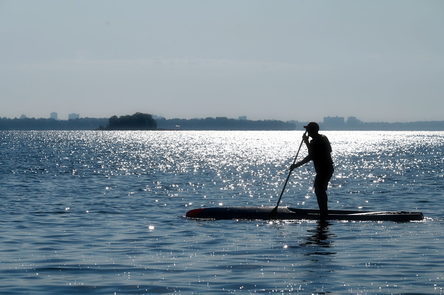 SUP Paddler paddles showing a sihlouette with shimmering water and Aylmer Island in the background.